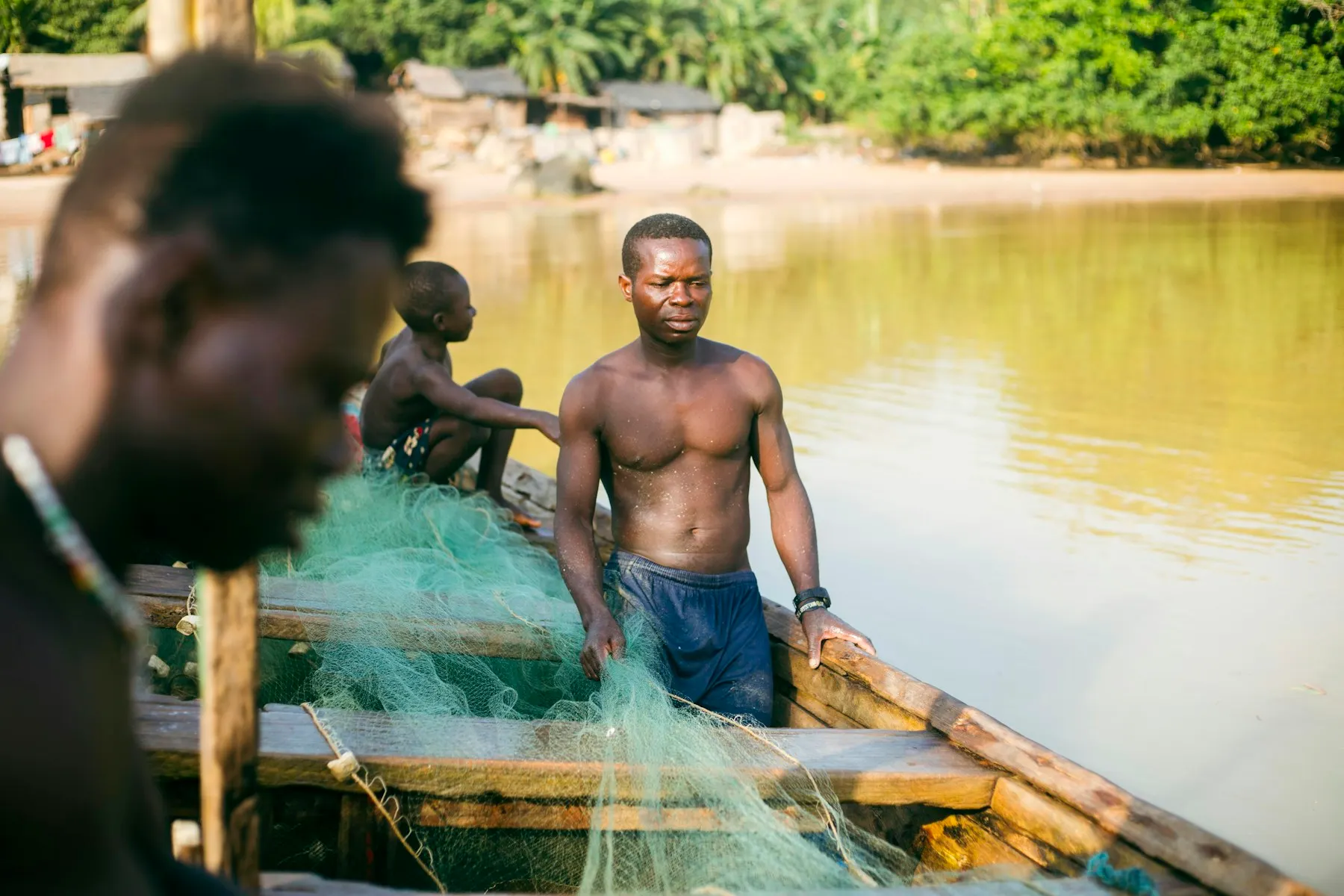 A fisherman sitting in a wooden boat with green nets beside a river