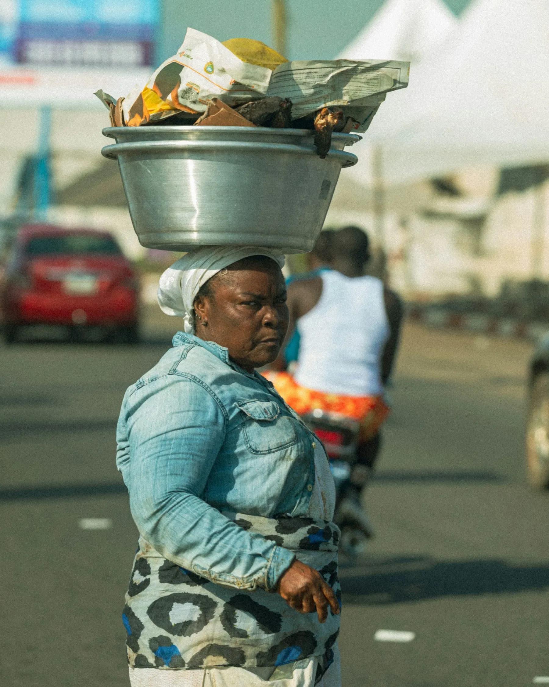 A woman carrying a large metal bowl of goods on her head along a road